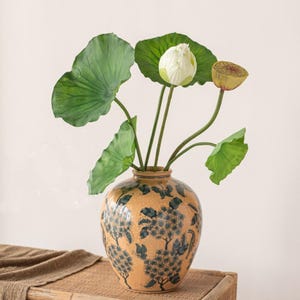 Könnte beinhalten: Eine Keramikvase mit floralem Design enthält Lotusblätter, eine weiße Lotusblüte und eine Samenkapsel. Die Vase hat einen beigefarbenen Hintergrund mit blauen Blumenmustern. Die Anordnung steht auf einer gewebten Oberfläche.