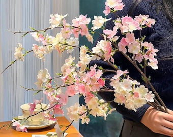 May include: Branches of artificial cherry blossoms with pink and white flowers. The branches are held by a person wearing a navy blue sweater. A teacup and saucer are on a wooden table.