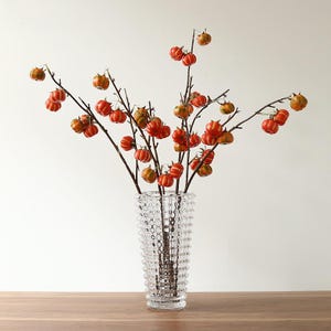 Puede incluir: Un jarrón de vidrio transparente con superficie texturizada sostiene ramas adornadas con pequeños adornos en forma de calabaza naranja y roja. El arreglo está sobre una superficie de madera sobre un fondo blanco, creando un ambiente otoñal.