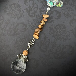 May include: A crystal suncatcher with a silver angel charm, brown stone beads, and a clear crystal prism. The suncatcher is hanging from a silver chain.