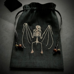 以下が含まれることがあります： 白い刺繍の蝙蝠の骨格のデザインが施された黒い巾着袋。袋には紐に茶色の木製トグルが付いています。