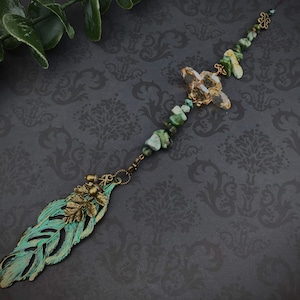 May include: A long, beaded necklace with a green and brown feather charm. The necklace features a variety of green and brown stones, as well as a clear crystal. The feather charm is made of metal and has a rustic, antique finish.