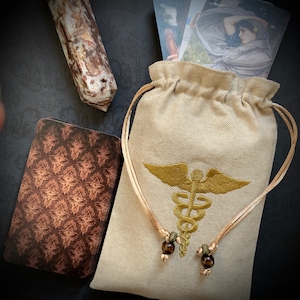 May include: A beige fabric pouch with a drawstring closure and a gold embroidered medical symbol. The pouch is sitting on a patterned surface with a brown and white stone object in the background.