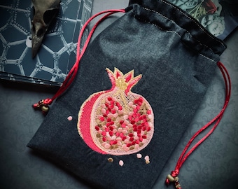 Könnte beinhalten: Eine schwarze Kordeltasche mit einem roten Kordelverschluss. Die Tasche hat ein gesticktes Granatapfel-Design mit goldenen, rosa und roten Akzenten.