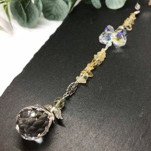 May include: A crystal suncatcher with a clear crystal ball at the bottom, a silver angel wing charm, and a silver flower charm. The suncatcher is strung with clear and yellow crystals.