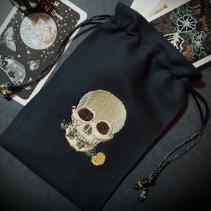 以下が含まれることがあります： 黒い紐で絞るタイプの袋で、前面に刺繍された骸骨が描かれています。骸骨は白く、黒い目と口の中に1つの黄色い花があります。袋は柔らかく黒い布でできています。