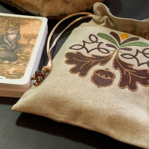 以下が含まれることがあります： オークの葉とドングリが茶色で刺繍されたベージュの巾着袋。袋には茶色と赤のビーズの紐が付いています。