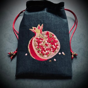 May include: A black drawstring bag with a red embroidered pomegranate design. The pomegranate is filled with pink and gold details.