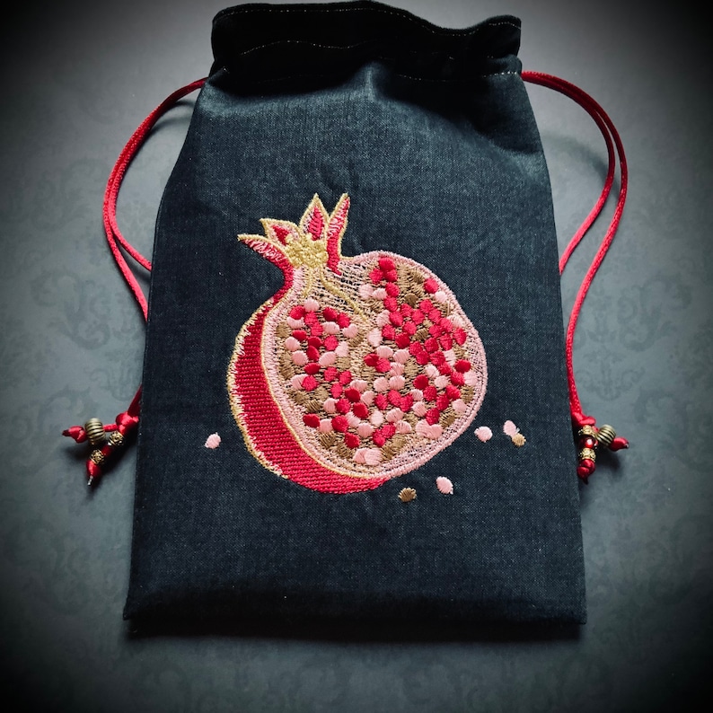 May include: A black drawstring bag with a red embroidered pomegranate design. The pomegranate is filled with pink and gold details.