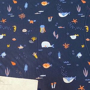 Könnte beinhalten: Ein dunkelblauer Stoff mit einem sich wiederholenden Muster von Meeresbewohnern, darunter Fische, Wale, Seepferdchen, Quallen und Seesterne. Das Muster ist in Blautönen, Orange und Gelb gehalten.