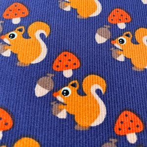 Könnte beinhalten: Ein blauer Stoff mit einem sich wiederholenden Muster aus orangefarbenen Eichhörnchen, die Eicheln halten, und roten Pilzen mit weißen Punkten.