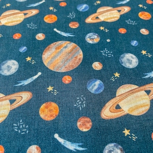 Könnte beinhalten: Ein blaugrüner Stoff mit einem sich wiederholenden Muster aus Planeten, Sternen und Kometen. Die Planeten sind in Orange-, Braun- und Blautönen gehalten. Die Sterne sind gelb und die Kometen sind blau.