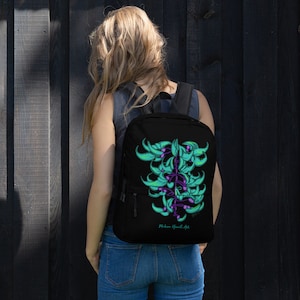 Puede incluir: Mochila negra con un diseño floral colorido en la parte posterior. El diseño presenta flores verdes y moradas con un contorno negro. El texto "Mokena Hawaii Art" está impreso en la parte inferior de la mochila.