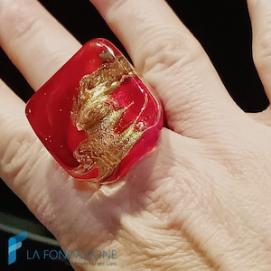 May include: A red glass ring with gold flecks. The ring is square shaped and has a unique, abstract design.