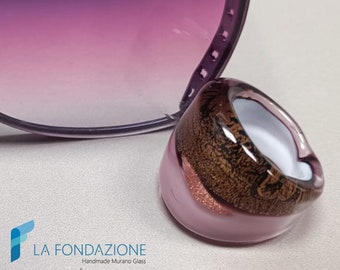 May include: A pink Murano glass ring with a brown and gold design, next to a pair of sunglasses with purple gradient lenses. The ring has a unique shape and a white interior. The sunglasses have a silver frame. The image includes the text "MADE IN ITALY".
