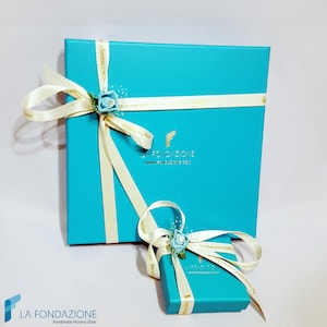 Puede incluir: Dos cajas de regalo turquesa con cinta de sat&eacute;n blanca y una flor azul. La caja m&aacute;s grande tiene el texto "LA FONDAZIONE www.lafondazione.com" impreso en ella.