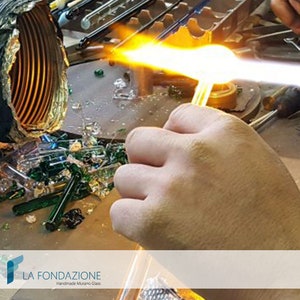 May include: A hand holds a glass rod over a flame, melting the glass. The glass is being heated to create a piece of Murano glass. The image includes the text "LA FONDAZIONE Handmade Murano Glass".