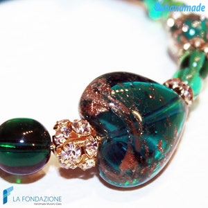 May include: A green glass heart charm with gold flecks and a gold and crystal bead. The charm is part of a bracelet with green glass beads.