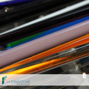 May include: Close-up of colorful glass rods, arranged in a stack. The rods are various shades of blue, black, white, orange, and purple. The rods are translucent and have a glossy finish. The text "LA FONDAZIONE Handmade Murano Glass" is visible at the bottom of the image.