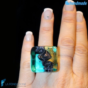 May include: Square ring made of colorful glass with swirls of black, turquoise, and gold. The ring is on a finger.