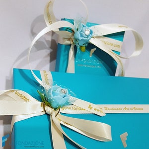 Puede incluir: Dos cajas de regalo turquesa con cinta de sat&eacute;n blanca y una flor azul claro con un centro de strass. Las cajas est&aacute;n envueltas con una cinta blanca con letras doradas que dicen "FONDAZIONE Handmade Murano Glass" y "www.fondazione.com Handmade Art in Venice".