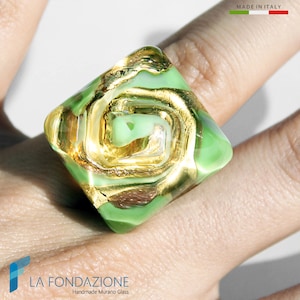May include: A square-shaped ring made of green Murano glass with gold leaf accents. The ring is on a finger.