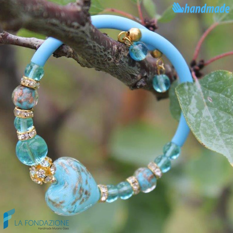 May include: A turquoise blue bracelet with a heart-shaped Murano glass charm. The bracelet is made with small turquoise blue glass beads and clear rhinestone accents. The charm has gold flecks and is surrounded by clear rhinestones. The bracelet is handmade and is on a branch with green leaves.