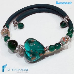 May include: A green heart-shaped Murano glass charm with gold flecks is the centerpiece of this bracelet. The bracelet is made with green and clear glass beads, silver spacers, and a dark green elastic band. The bracelet is handmade and features the text "LA FONDAZIONE Handmade Murano Glass".