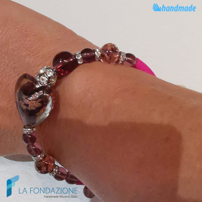 May include: A pink elastic bracelet with a heart-shaped Murano glass charm. The charm is a deep burgundy color with gold flecks and clear glass accents. The bracelet is adorned with smaller burgundy and clear glass beads and silver spacers.
