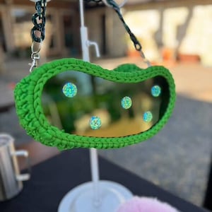 Op de afbeelding: Een groene gehaakte tas met een doorzichtig midden, versierd met iriserende versieringen. De tas heeft een donkergroene kettingriem en wordt tentoongesteld op een witte standaard. Een roze, pluizig accessoire is hieronder zichtbaar.