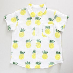 Könnte beinhalten: Weißes kurzärmeliges Hemd mit gelbem Ananasmuster. Das Hemd hat einen Kragen, eine Vordertasche und Knopfverschlüsse. Die Ananas sind gelb mit grünen Spitzen.