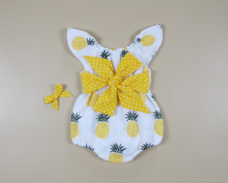 May include: White baby romper with yellow pineapple print and a large yellow polka dot bow. A matching yellow bow is also included. The romper has flutter sleeves and is designed for infants.