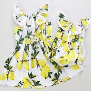 May include: Two white dresses with yellow lemons printed on them. The dresses have white ruffled collars and white pom-pom trim.