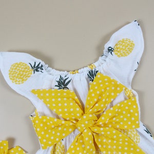May include: White baby dress with yellow pineapple print and a large yellow polka dot bow. The dress has ruffled sleeves and a gathered neckline. The overall design is summery and playful.