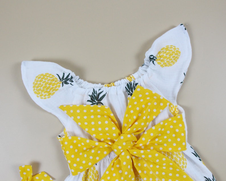 May include: White baby dress with yellow pineapple print and a large yellow polka dot bow. The dress has ruffled sleeves and a gathered neckline. The overall design is summery and playful.