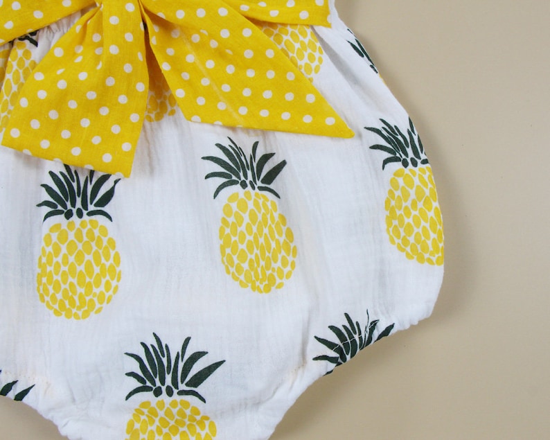 May include: White romper with a yellow pineapple print and a large yellow bow with white polka dots. The romper features a playful design with a tropical theme, perfect for warm weather.
