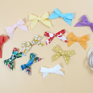 May include: A collection of colorful fabric bows in various shades of pink, yellow, blue, purple, red, and white. Some bows feature floral patterns. Several neutral-colored headbands are also visible, suggesting a hair accessory theme.