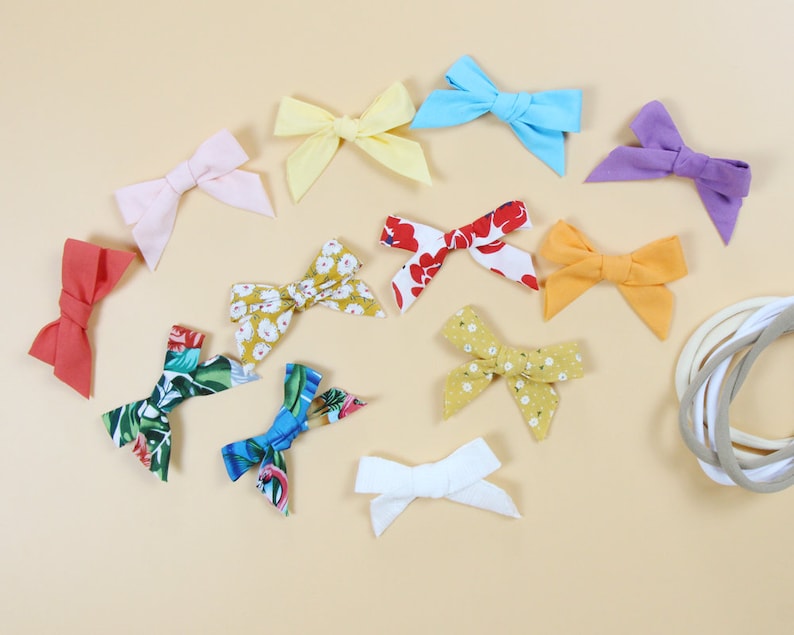 May include: A collection of colorful fabric bows in various shades of pink, yellow, blue, purple, red, and white. Some bows feature floral patterns. Several neutral-colored headbands are also visible, suggesting a hair accessory theme.
