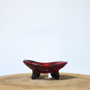 Könnte beinhalten: Eine kleine, flache Keramikschale mit einer glänzenden, gesprenkelten rot-schwarzen Glasur. Die Schale hat einen geschwungenen, organischen Rand und wird von drei kurzen, stabilen, mattschwarzen Beinen getragen, die den Boden von der Unterlage abheben.