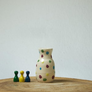 Könnte beinhalten: Eine kleine, cremefarbene Keramikvase mit bauchigem Körper und engem Hals, verziert mit verstreuten Punkten in Blau, Gelb, Grün und Kastanienbraun. Neben der Vase stehen drei hölzerne Spielfiguren in den Farben Grün, Gelb und Blau.
