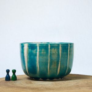 Könnte beinhalten: Eine Keramikschale mit glänzender türkisfarbener Glasur und vertikalen, weißen, tropfenartigen Streifen. Neben der Schale befinden sich zwei kleine, glatte Spielfiguren, eine in Blau und eine in Grün, auf einer runden Holzoberfläche.
