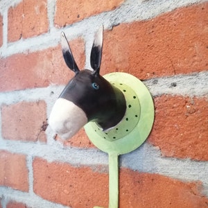 Puede incluir: Un gancho de pared decorativo con forma de cabeza de burro. La cabeza del burro es negra y blanca con ojos azules, montada sobre una base circular verde claro con puntos negros. El gancho es de metal verde claro curvado.