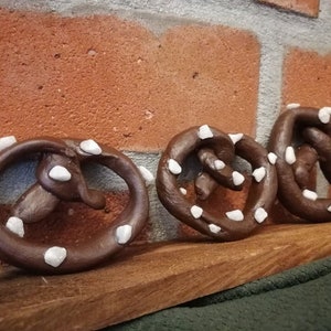 May include: Three brown pretzel shaped ornaments with white decorative accents. The ornaments are arranged on a wooden surface.