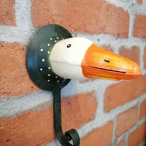 Może przedstawiać: Kapryśny wieszak ścienny w kształcie głowy gęsi. Gęś ma białą głowę, pomarańczowy dziób i czarne oko. Hak jest przymocowany do ciemnozielonej okrągłej podstawy z białymi kropkami i czarnym, zakrzywionym metalowym hakiem.