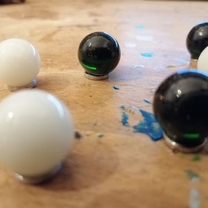 K&ouml;nnte beinhalten: Eine Ansammlung von Glasmurmeln, einige wei&szlig; und einige schwarz, die auf einer Holzoberfl&auml;che liegen. Jede Murmel hat eine gl&auml;nzende, reflektierende Oberfl&auml;che. Die schwarzen Murmeln haben einen gr&uuml;nen Streifen. Die Murmeln stehen auf kleinen, silberfarbenen Sockeln.