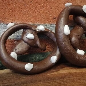 May include: Two brown pretzel-shaped ornaments decorated with white stones. The ornaments are sitting on a wooden surface.