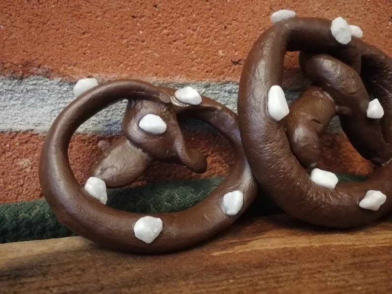May include: Two brown pretzel-shaped ornaments decorated with white stones. The ornaments are sitting on a wooden surface.