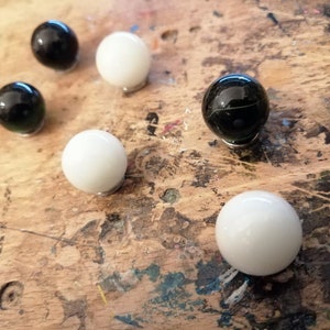 K&ouml;nnte beinhalten: Eine Ansammlung von Murmeln, einige schwarz und einige wei&szlig;, liegt auf einer Holzoberfl&auml;che. Die Murmeln sind gl&auml;nzend und kugelf&ouml;rmig, mit reflektierenden Eigenschaften. Die Holzoberfl&auml;che hat ein abgenutztes Aussehen.