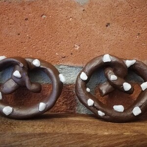 May include: Three brown pretzel-shaped ornaments with white decorative accents. The ornaments are made of a smooth, dark brown material and have a slightly glossy finish. Each ornament has a different arrangement of white accents, which are small and round.