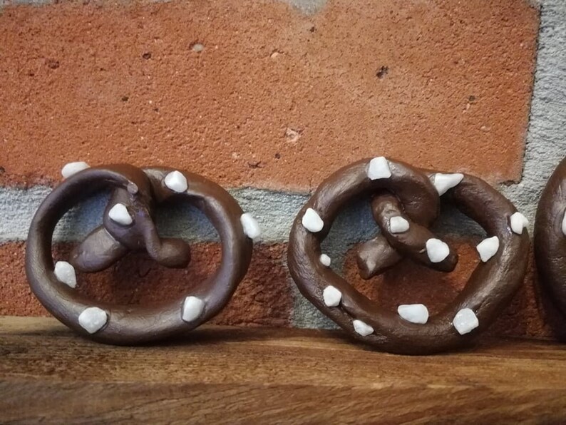 May include: Three brown pretzel-shaped ornaments with white decorative accents. The ornaments are made of a smooth, dark brown material and have a slightly glossy finish. Each ornament has a different arrangement of white accents, which are small and round.